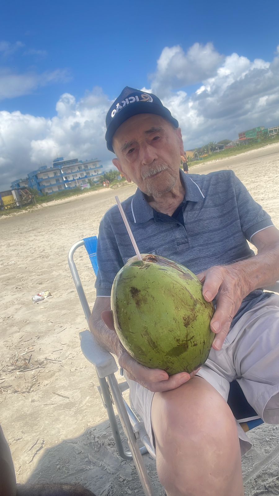 Senhor Pedro Sandoli, sentado em cadeira na praia segurando um coco verde, usando boné